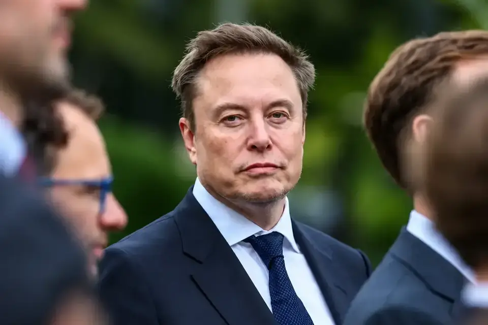 Elon Musk in a dark suit and tie standing among other people outdoors, in coverage of his reported plan to give SpaceX shares to children.