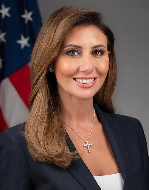 A woman in a white blazer, her hair in a long ponytail, standing in front of the U.S. Capitol.